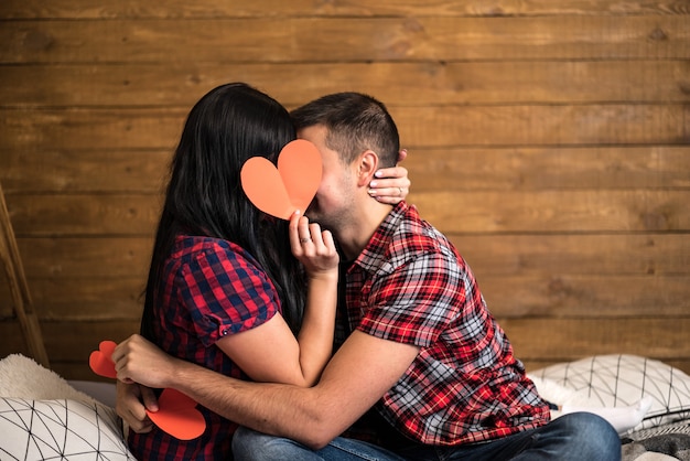 Romántica pareja sentada en un sofá en un amoroso abrazo besándose mientras sostienen un corazón de papel rojo sobre madera