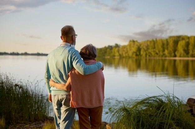 Romántica y tranquila pareja de ancianos mirando desde lejos de pie en la orilla del río