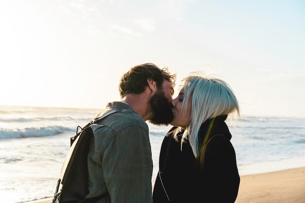 Romântico casal feliz apaixonado beijando no litoral na praia selvagem Namorado e namorada vestindo roupas legais e mochila vintage em frente ao mar durante o pôr do sol