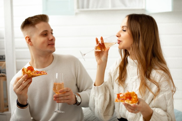 Romántico hombre y mujer de pelo rubio vestidos de blanco, celebrando alguna festividad posiblemente el día de san valentín, sonriendo, bebiendo champán en copas, comiendo pizza y mirándose el uno al otro