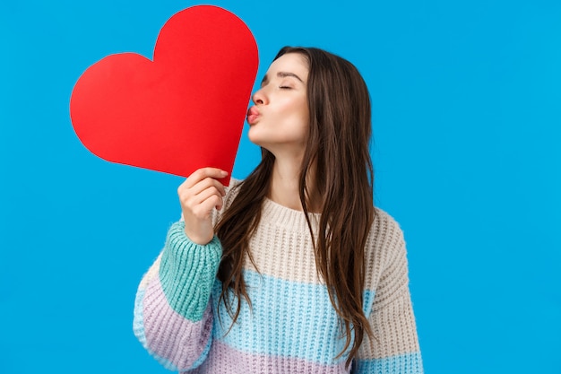 Romantik, Liebe und Zärtlichkeit Konzept. Weibliche fröhliche und niedliche brünette Frau zeigt ihre Gefühle, küsst große rote Herzkarte mit geschlossenen Augen, drückt Sympathie und Fürsorge aus, blaue Wand