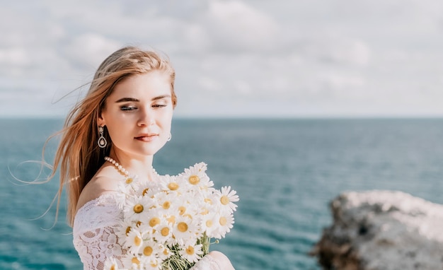 Romantische Braut blondes Mädchen im weißen Hochzeitskleid mit offenen Schultern posiert mit Meer und Felsen im Hintergrund Stilvolle junge Frau, die am Rand einer Klippe steht und bei Sonnenuntergang in die Berge eines Ozeans blickt