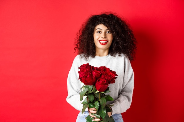 Romantische Frau mit lockiger Frisur erhalten Blumenstrauß von roten Rosen am Valentinstag, zärtlich auf Liebhaber schauend, stehend auf Studiohintergrund.