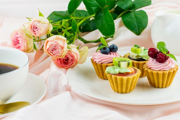 Romantische Komposition für Valentinstag, Urlaub, Geburtstag. Mini-Törtchen mit Sahne und Früchten auf einem Teller, eine Tasse Kaffee, ein Rosenstrauß.