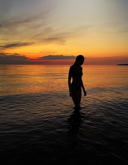 romantische Silhouette der jungen Frau bei orangefarbenem Sonnenuntergang in der Meerwasserwelle und rosa bewölktem Himmel