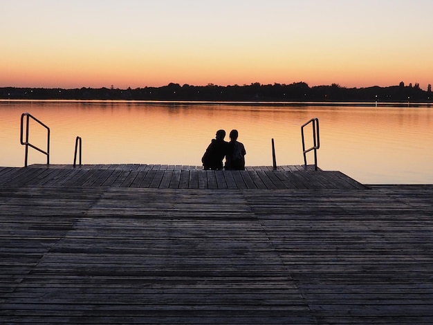 Romantische Szene Junges Paar sitzt auf dem Pier am See bei Sonnenuntergang Ein Mann und eine Frau umarmen sich Blick auf die Sonne Rückansicht Liebe und Datum Valentinstag See Palich Serbien Oranger Himmel