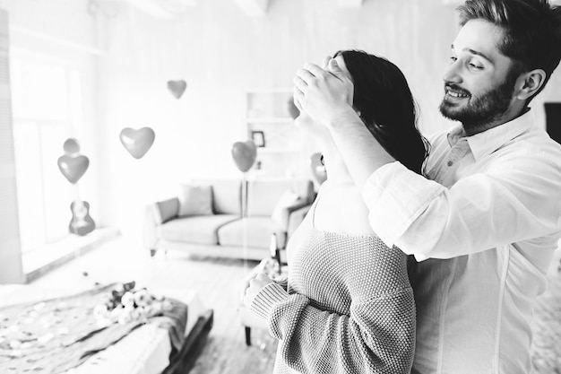 Romantische überraschung für ihre geliebte. ein gutaussehender mann umarmt und schließt die augen zu seiner freundin oder seiner frau und macht sie zu einem romantischen abendessen im schlafzimmer für ihren jahrestag oder valentinstag