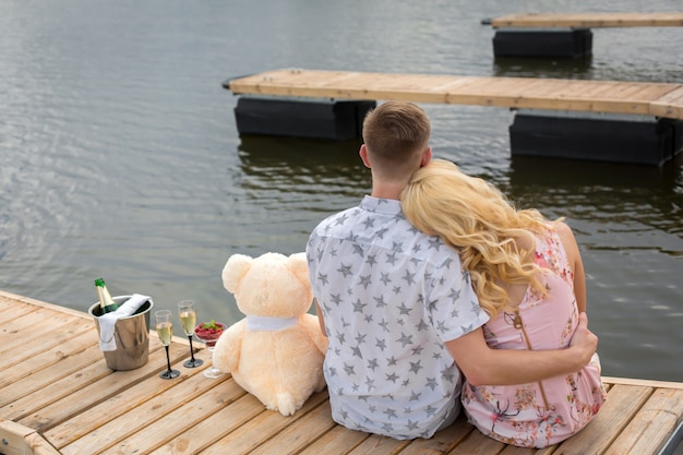 Foto romantische verabredungsüberraschung, ein junger mann und ein mädchen auf einem holzsteg, umarmen und küssen beim sitzen auf dem steg, romantische liebesgeschichte