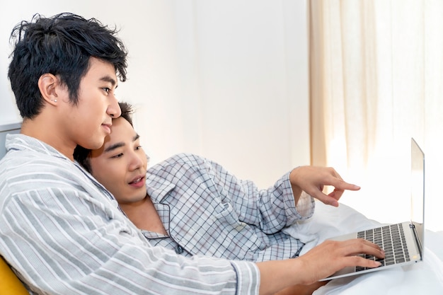 Romantische Zeit. Asiatische homosexuelle Paare in den Pyjamas online kaufend mit Computernotizbuch im Bett. Konzept LGBT Homosexuell.