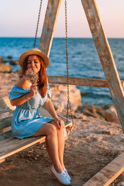 Romantischer Abend bei Sonnenuntergang am Meer, Frau mit Blumen sitzt auf einer hölzernen Schaukel, die Hut hält