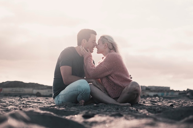 Romantischer Abend mit Mädchen und Jungen, die zusammen am Strand auf dem Sand sitzen und sich in Zärtlichkeit küssen Zwei Personen, männlich und weiblich, in Beziehung, die auf dem Sand sitzen und den Sonnenuntergang genießen Mann, Frau, Kuss