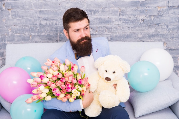 Romantischer Mann mit Blumen und Teddybär sitzen auf der Couch mit Luftballons wartende Freundin Romantisches Geschenk Macho bereit romantisches Date Mann trägt Smoking Fliege halten Blumenstrauß Sie verdient alles Gute