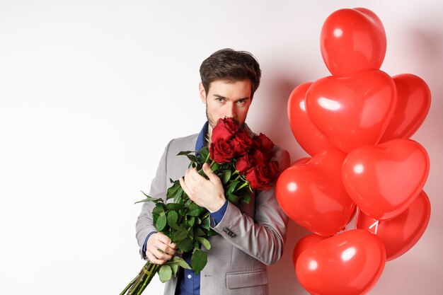 Romantischer Mann riecht Strauß roter Rosen und schaut leidenschaftlich in die Kamera. Freund im Anzug, der auf Valentinstag mit Geschenken und Herzballons, weißer Hintergrund geht.