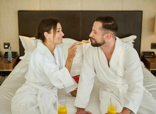 Romantischer Morgen eines jungen schönen Liebespaares, das das Frühstück im Bett für zwei genießt. Frischvermählte füttern sich gegenseitig leckeres Gebäck, während sie während ihrer Flitterwochen im Bett in einem Hotelzimmer frühstücken