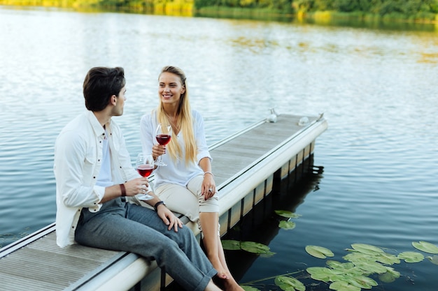 Romantischer Ort. Erfreut freudiges Paar, das in der Nähe des Flusses sitzt und seinen Wein genießt