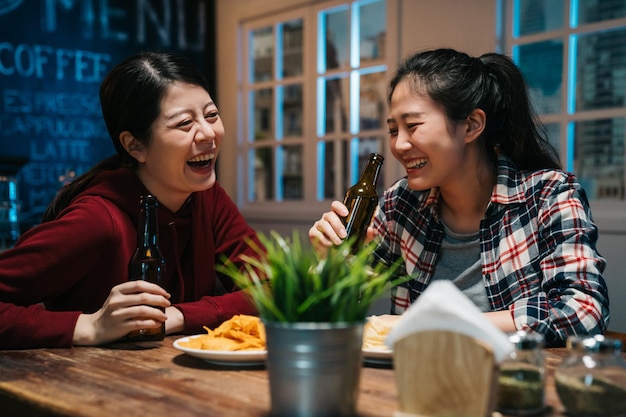 romantisches asiatisches lesbisches paar, das bier in der flasche in der vintage-bar in der modernen stadt trinkt. Junge Mädchen, die in der Damennacht im Pub lachen, genießen Alkohol und betrinken sich am Holztisch mit Pommes.