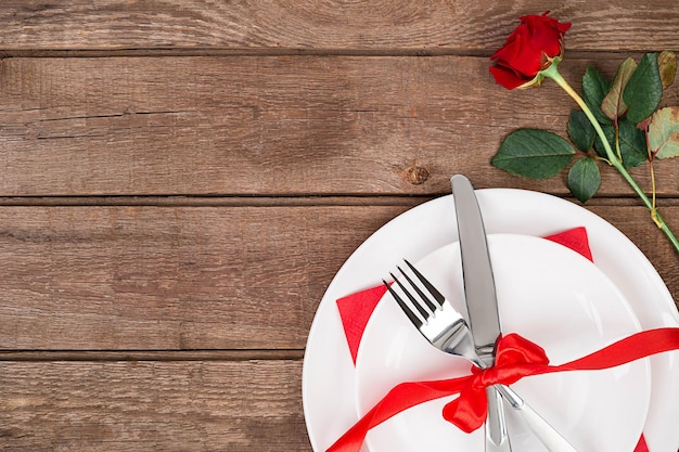 Romantisches Dinner-Konzept. Valentinstag oder Vorschlagshintergrund. Blick von oben auf den Holztisch des Restaurants mit Herz und Rose mit Besteck auf dem Teller. Kopieren Sie Platz auf rustikalem Holz