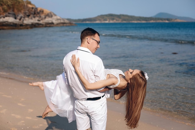 Romantisches Foto eines Hochzeitstages. Schöner Ehemann, der seine junge schöne Frau in einem Hochzeitskleid an der Küste in seinen Armen hält. Konzept der Hochzeit.