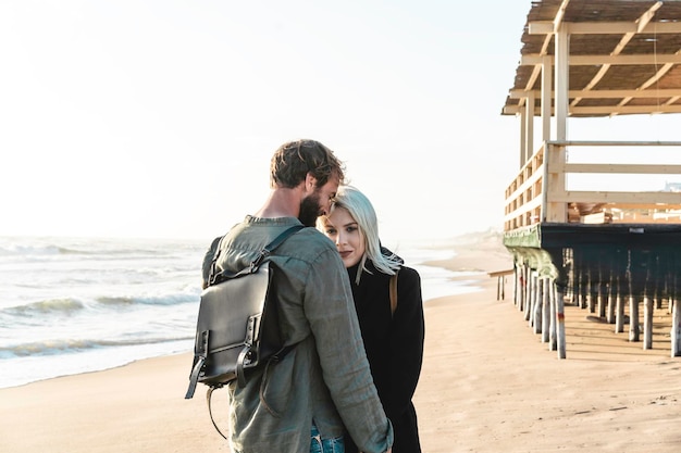 Romantisches Hipster-Paar in Liebe, das an der Küste am wilden Strand in der Nähe der Holzstruktur steht Freund und Freundin tragen coole Kleidung und einen Vintage-Rucksack, der vor dem Meer steht