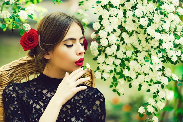 Romantisches Mädchen oder hübsche Frau mit weißer Blume im Mund mit roten Lippen stilvolles Make-up auf nachdenklichem Gesicht und Rosen im brünetten Haar auf floraler Umgebung Frühling und Sommer Schönheit der Natur