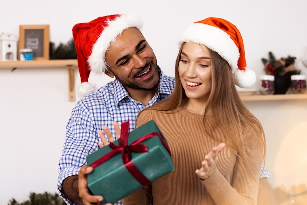 Romantisches multiethnisches Paar in Weihnachtsmützen, die Geschenke austauschen