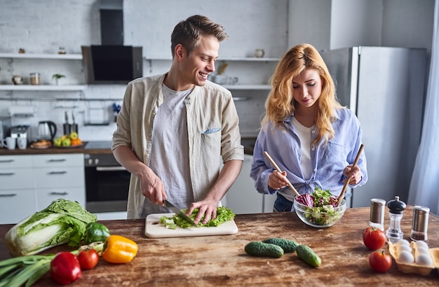 Romantisches Paar kocht in der Küche. Gut aussehender Mann und attraktive junge Frau haben Spaß zusammen, während sie Salat machen. Gesundes Lebensstilkonzept.