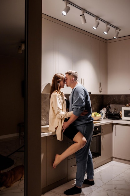Romantisches, sexy junges Paar, das glücklich Zeit damit verbringt, sich in einer gemütlichen, modernen Küche zu Hause zu umarmen und zu küssen Zwei Personen stehen und sehen sich freudig an