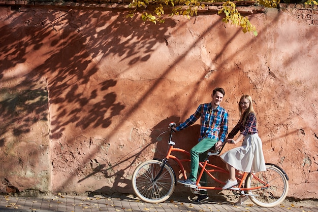 Romantisches Touristenpaar, bärtiger Mann und blonde Frau, die zusammen stehen und zur Kamera auf Doppel-Tandemfahrrad auf hoher roter alter knisternder verputzter Wand lächeln.