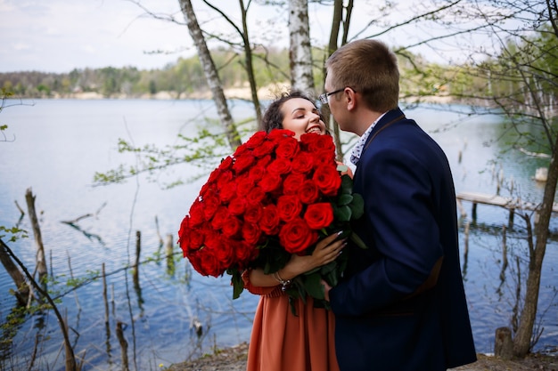 Romantisches treffen junger leute. ein mann im anzug mit einem strauß roter rosen gibt dem mädchen einen strauß und sie küssen sich im wald