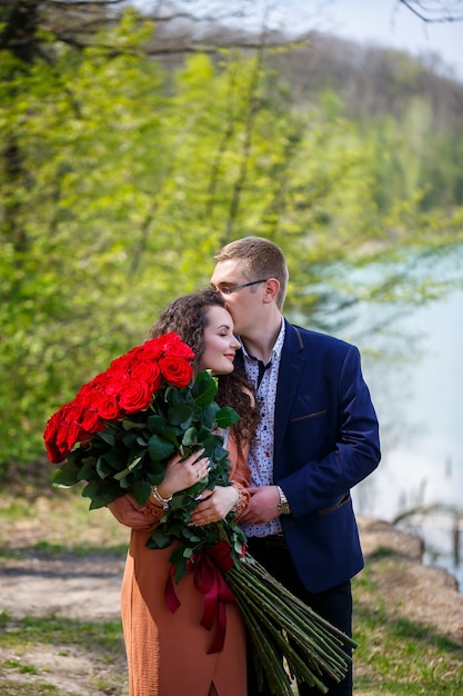 Romantisches Treffen junger Leute. Eine junge Frau erklärte sich bereit, ihren Mann zu heiraten. Ein Mann im Anzug mit einem Strauß roter Rosen gibt dem Mädchen einen Strauß und sie küssen sich im Wald