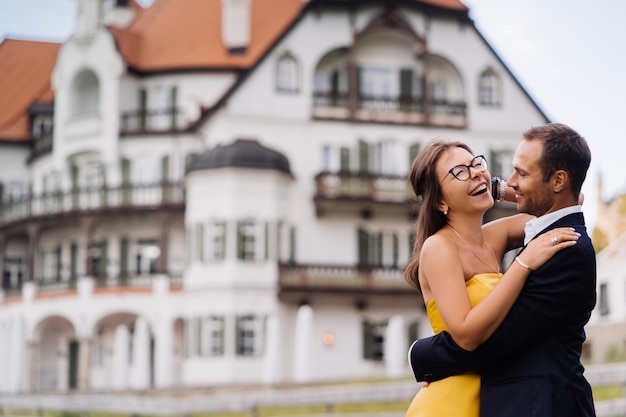 Romantisches verliebtes Paar, das gegen altes Gebäude umarmt. Junge Frau im gelben Abendkleid, glücklich lachend, nachdem sie eine gute Nachricht erhalten hat. Konzept der glücklichen Leute.