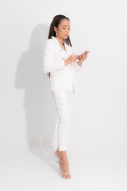 Ropa de catálogo de estilo de moda para mujer de negocios, cabello largo negro, maquillaje natural, traje de traje blanco, traje de forma de cuerpo perfecto, usando el teléfono móvil en la sesión de estudio sobre fondo blanco y sombra.