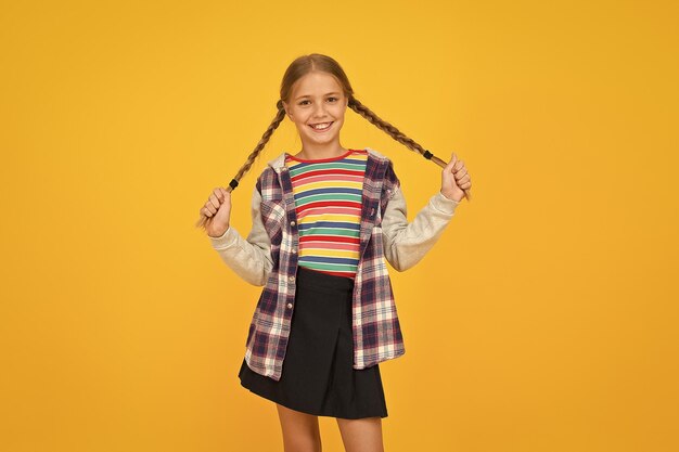 Ropa colorida para niña preadolescente Tienda para niños Centro comercial Chica adorable cara hermosa Peluquería Niña Niño pequeño con lindo peinado de trenzas sobre fondo amarillo Concepto de cuidado infantil