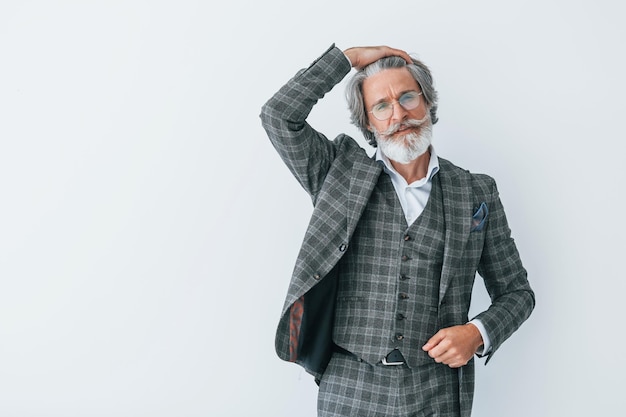 En ropa elegante De pie contra la pared blanca Hombre moderno con estilo senior con cabello gris y barba en el interior