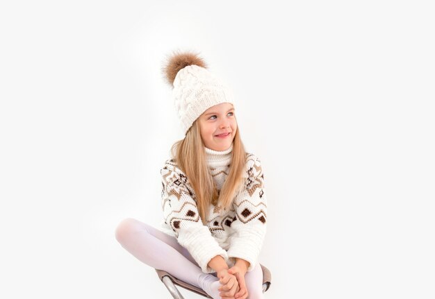 Ropa de invierno. Retrato de una niña de pelo rizado con un gorro de invierno blanco tejido. Niña rubia con gorro de punto blanco y un suéter sonriendo fondo claro aislar