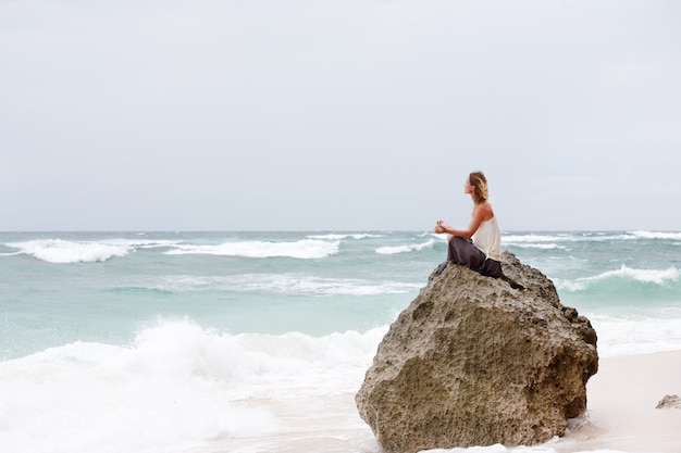 Ropa de niña hermosa en blanco sentarse en la orilla del mar en la roca y meditar en pose de mujer de yoga
