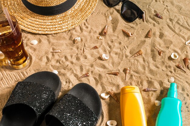 Ropa de playa de verano, chanclas, sombrero, bebida fría en un vaso y conchas marinas en la playa de arena