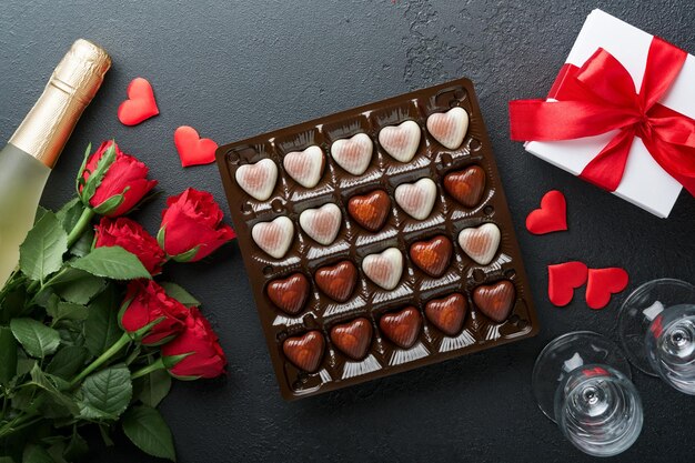 Rosa flores rojas galletas de brownie y dulces de chocolate en caja de regalo en forma de corazón con vino sobre fondo negro Día de San Valentín Tarjeta de felicitación del Día de la Madre o de la Mujer Vista superior plana con espacio de copia