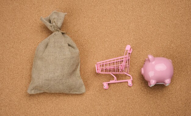 Foto rosa keramik-sparschwein, miniatur-einkaufswagen und voller canvas-tasche auf braunem hintergrund, ansicht von oben.