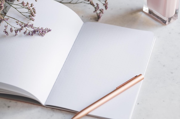 Rosa leeres Notizbuch aus Papier mit goldenem Stift auf weißem Tisch, umgeben von Blumen und Duft Das Konzept des weiblichen Arbeitsplatzes