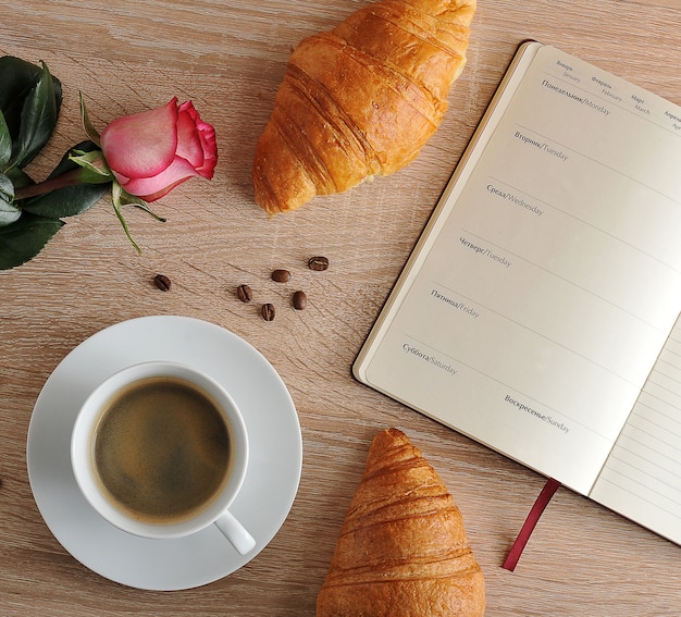 Rosa roja y una taza de café con un croissant y un diario con los días de la semana.