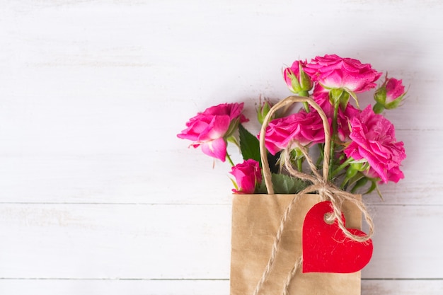 Rosa Rosen auf Bastelpapiertüte mit rotem Papierherz auf hölzernem weißem Hintergrund