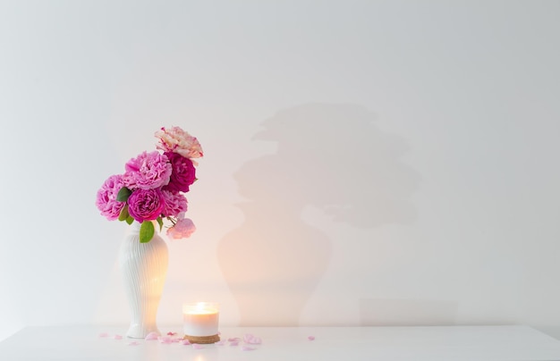 Rosa Rosen in Vase und brennende Kerze auf weißer Hintergrundwand