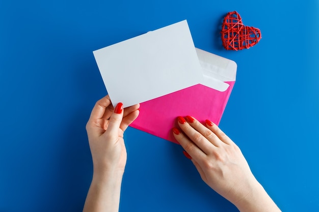 Rosa Umschlag mit einer leeren Karte in den Händen auf einem blauen Hintergrund. Umschlag mit einer sauberen Grußkarte und einem Herzen auf einem blauen Hintergrund. Konzept für Design am Valentinstag.