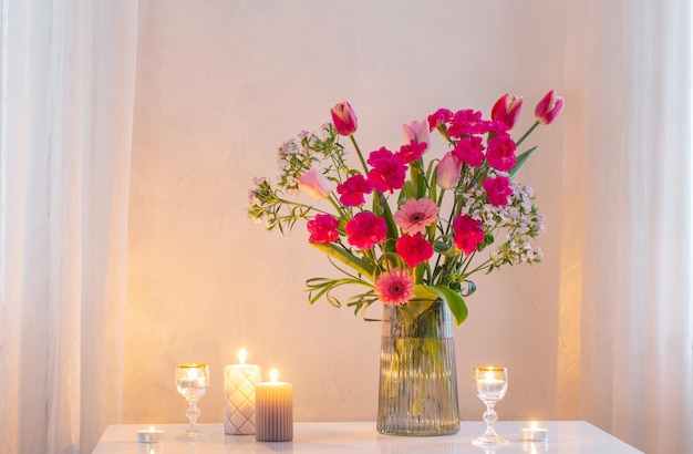 Rosafarbene Blumen in moderner Glasvase mit brennenden Kerzen im weißen Innenraum