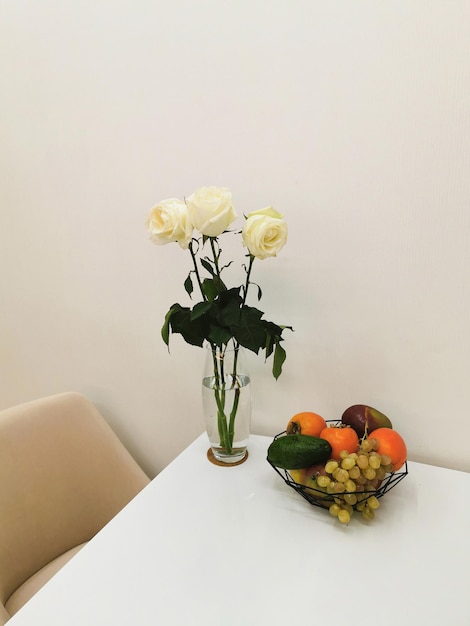 Foto rosas brancas em um vaso de vidro sobre uma mesa branca com um prato de frutas com uvas abacates