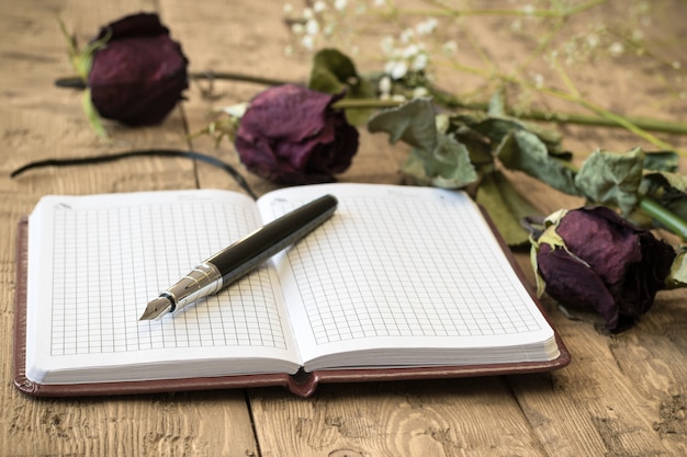 Rosas murchas com um caderno e uma caneta-tinteiro em uma mesa rústica.