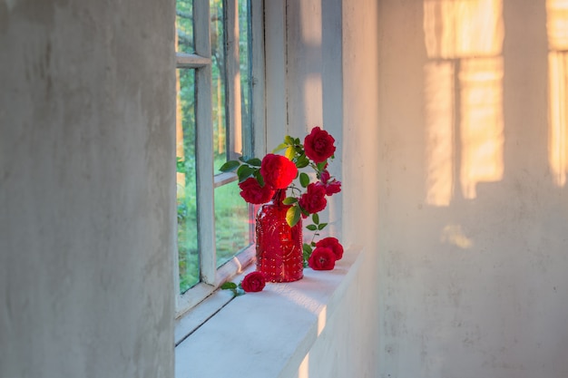 Rosas rojas en florero de cristal rojo en el alféizar de la ventana