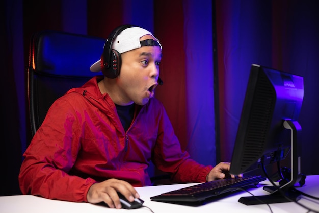 Rosto animado e chocado de um jogador asiático com fone de ouvido jogando videogame de computador online sentado na cadeira na sala de estar. Jogador profissional indiano transmitindo no jogo social muito divertido