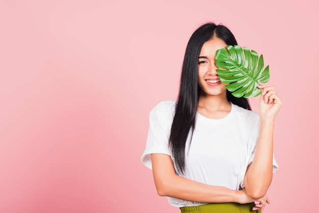 Rosto bonito. Retrato de uma bela jovem asiática com pele fresca e saudável segura folha de monstera verde em seu rosto, Folhas tropicais, foto de estúdio isolada em fundo rosa, Conceito de spa para cuidados com o corpo da pele